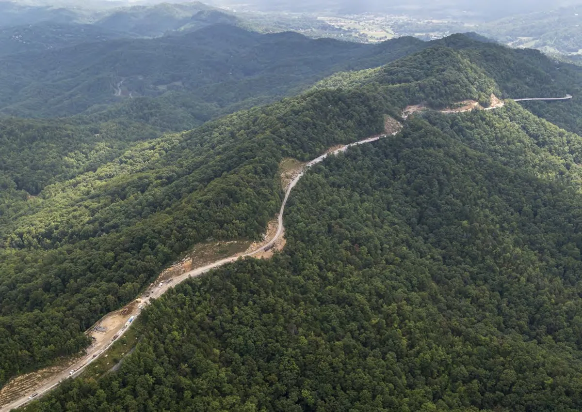 A breathtaking view of a mountain range stretches across the horizon, with a winding road cutting through dense forest and rolling hills. The natural landscape is vibrant and expansive, inviting adventure and exploration.