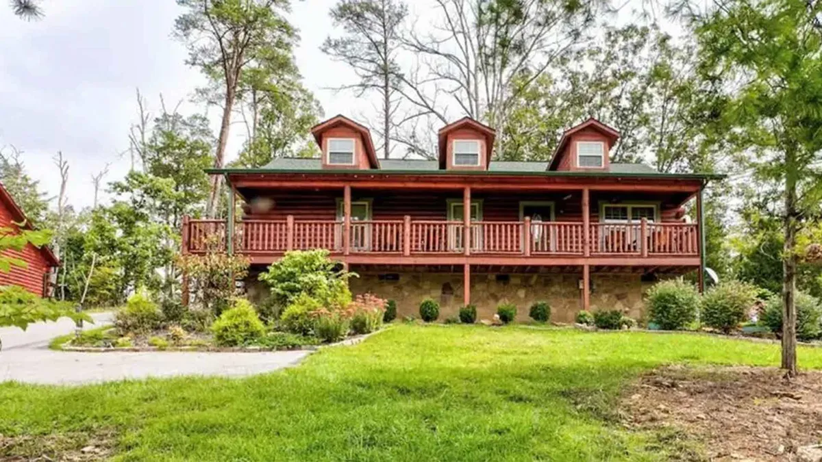 The image showcases the warm wooden exterior of a classic cabin with inviting triple dormer windows, a spacious porch perfect for gatherings, and lush landscaping all around. The porch offers stunning sunset views and a peaceful spot for morning coffee, surrounded by the natural beauty of Sevierville's rolling hills.