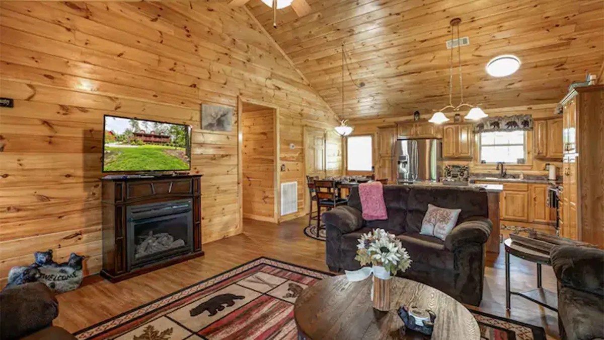 This warm and inviting shot captures the main living area, featuring a cozy fireplace, a television, a decorative rug, and an adjoining dining area next to wooden cabinets. Sunlight filters in through generous windows, creating a welcoming atmosphere perfect for gathering, relaxing, and entertaining.