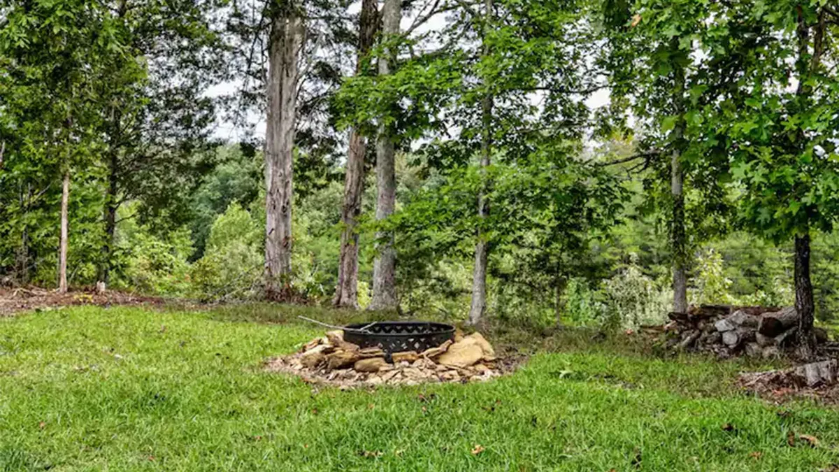 The image centers on a welcoming fire pit surrounded by green grass and tall trees, creating a cozy gathering spot. The fire pit is ready for an evening campfire, with plenty of space for friends or family to gather, roast marshmallows, and share stories under the stars.