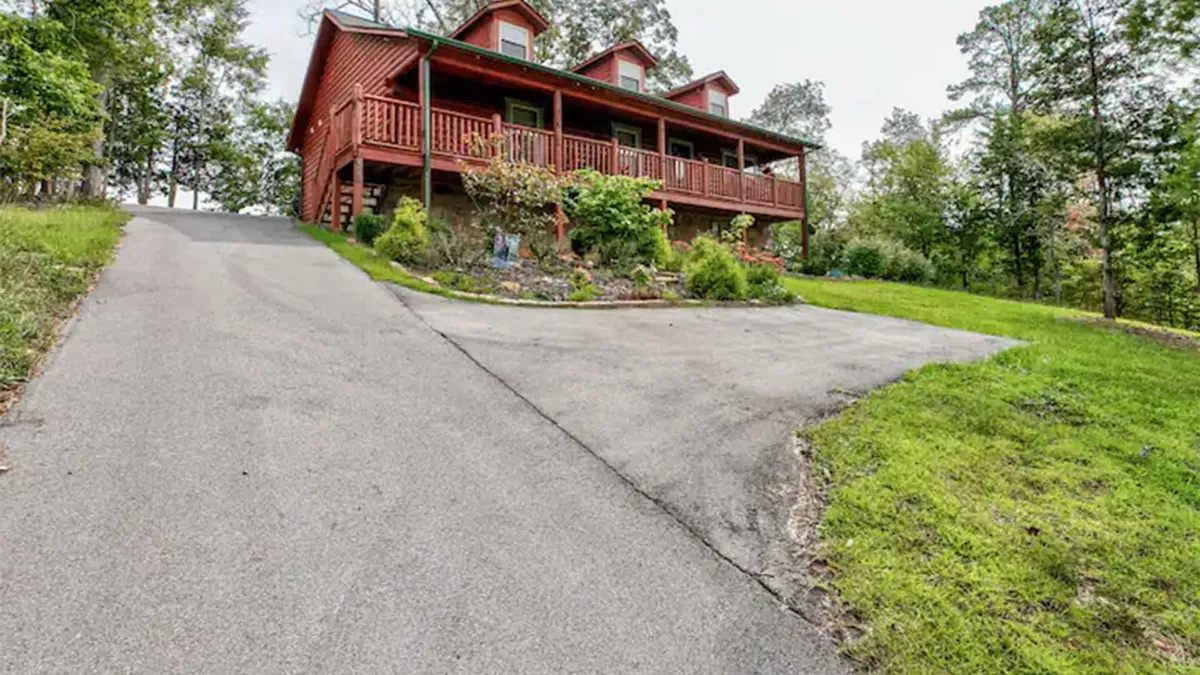 A charming red wooden house stands surrounded by lush landscaping, with a welcoming porch and rustic railing. The setting is tranquil and inviting, perfectly blending with the natural beauty of the area. Morning sunlight filters through the trees, casting gentle shadows across the garden and porch seating area.