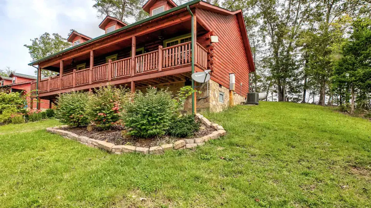 A welcoming wooden porch wraps around the cabin, surrounded by a lush garden and stone landscaping. The inviting outdoor space hints at cozy gatherings, relaxation, and enjoying nature together.