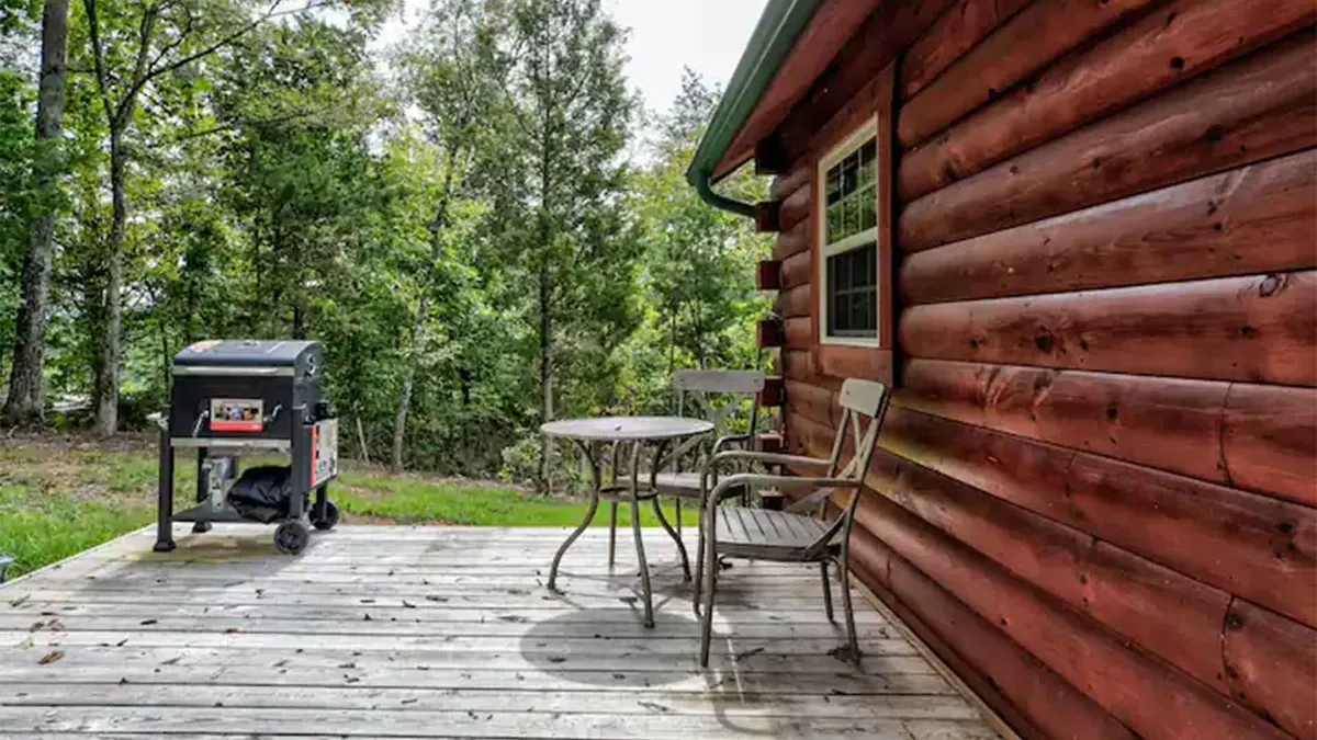 A wooden log cabin exterior features a spacious deck with outdoor seating and a grill, blending rustic charm with comfortable amenities. The deck is designed for both relaxation and social gatherings, with sunset lighting suggesting the perfect ambiance for evening conversations or early morning rituals.
