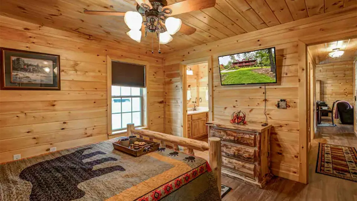 A cozy bedroom featuring a wooden bed frame with a bear-themed quilt, rustic dresser, wall-mounted TV, and a wooden ceiling. The room feels inviting and warm, with touches of mountain-themed decor, perfect for relaxing after a day of adventure.